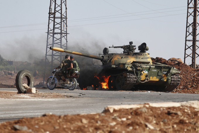Binh sĩ quân đội Syrian cưỡi xe máy đi qua một chiếc xe tăng bị bắn cháy ở ngoại ô thành phố Sweida ngày 14/7. Ảnh: AP